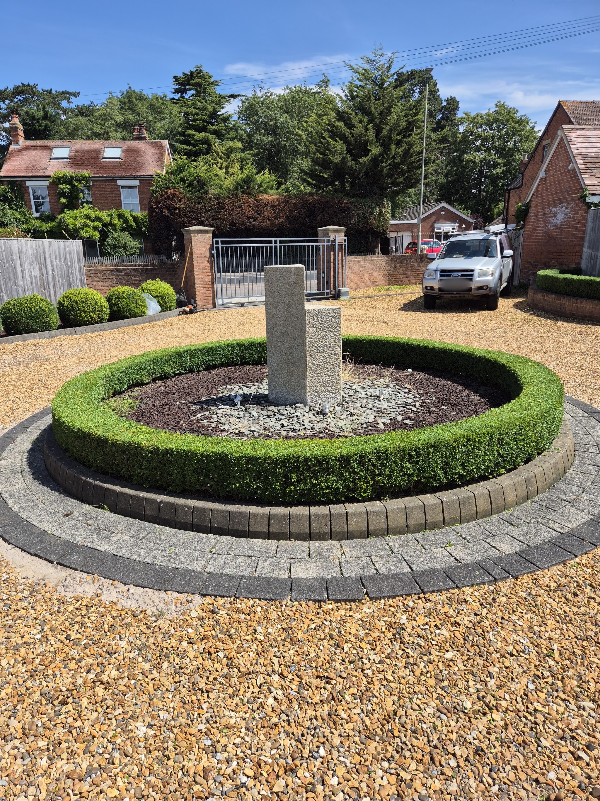 A circular box hedge clipped around a stone feature on a gravel drive